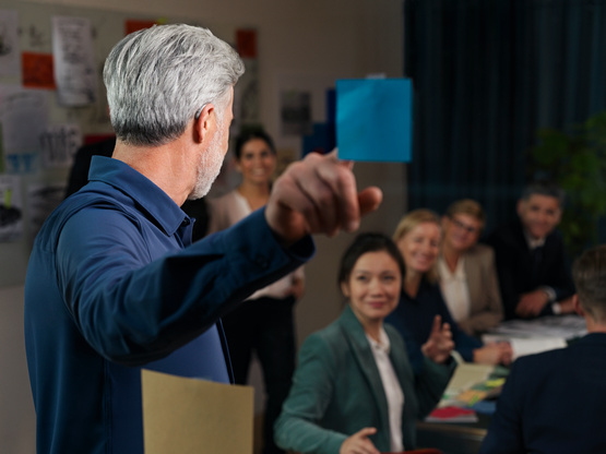 Munka értekezleten hallókészülékes férfi beszél a többiekhez és rámutat egy kék papírcetlire.